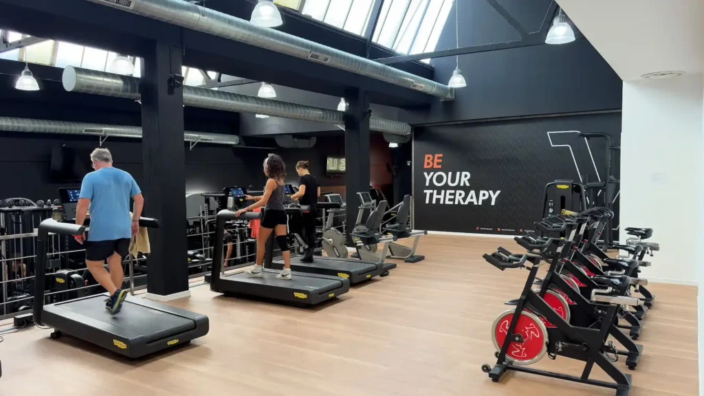 Ampia area cardio luminosa della palestra Vibe Fitness ad Arcore con persone che corrono sui tapis roulant, cyclette e scritta a parete Be Your Therapy
