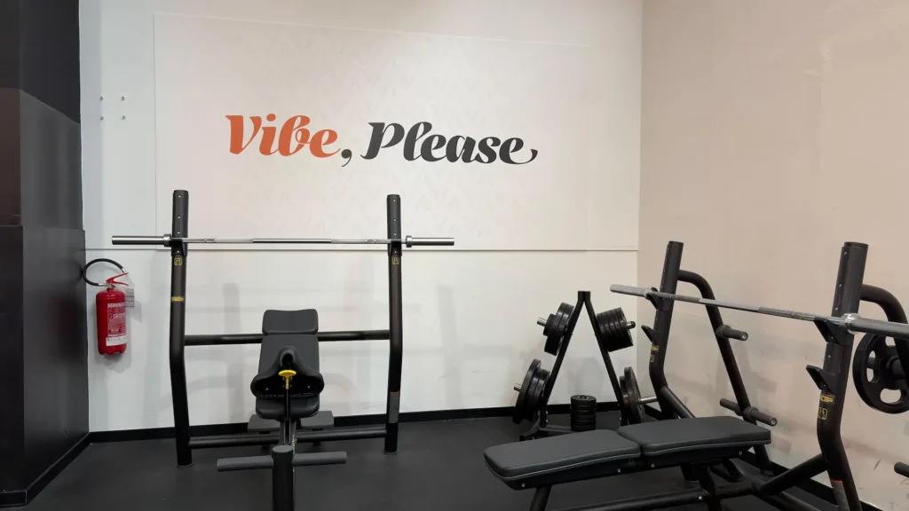 Postazione per panca piana con bilanciere e rastrelliera per dischi nella zona pesi liberi della palestra Vibe Fitness ad Arcore, sotto la scritta Vibe, Please