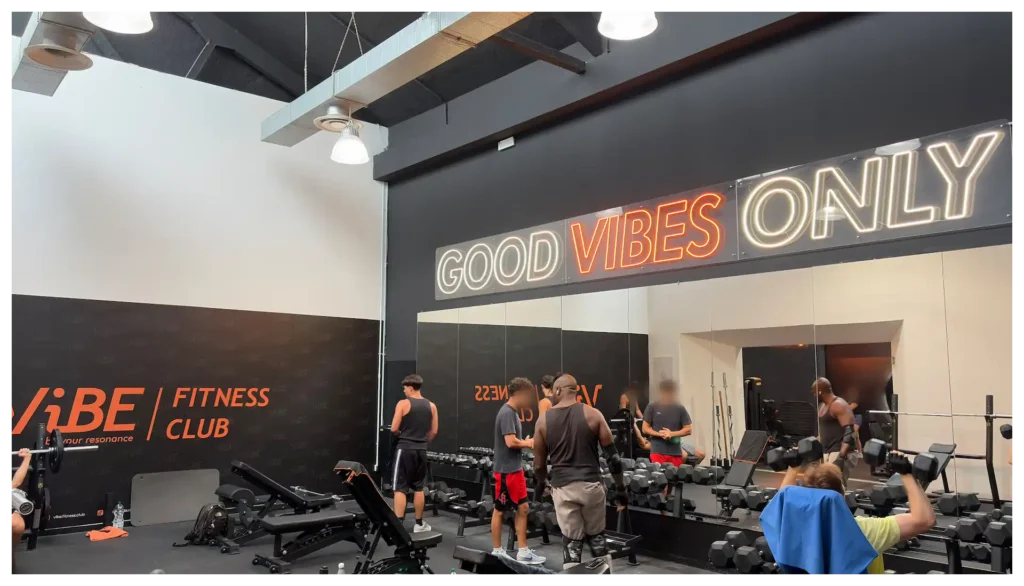 Sala pesi della palestra Vibe Fitness Club ad Arcore con persone che si allenano ai manubri e grande insegna luminosa con la scritta Good Vibes Only.