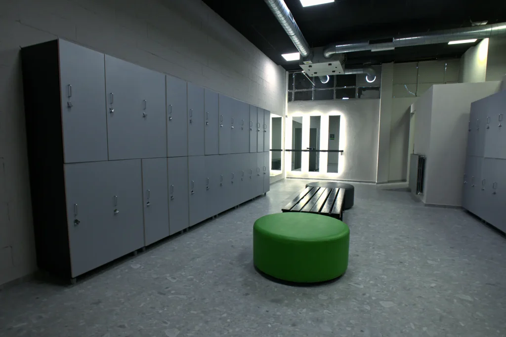 Vista interna degli spogliatoi della palestra ViBE Fitness ad Arosio, con file di armadietti grigi, una panca in legno e sedute pouf colorate (una verde brillante e una grigia) su un pavimento in terrazzo grigio.
