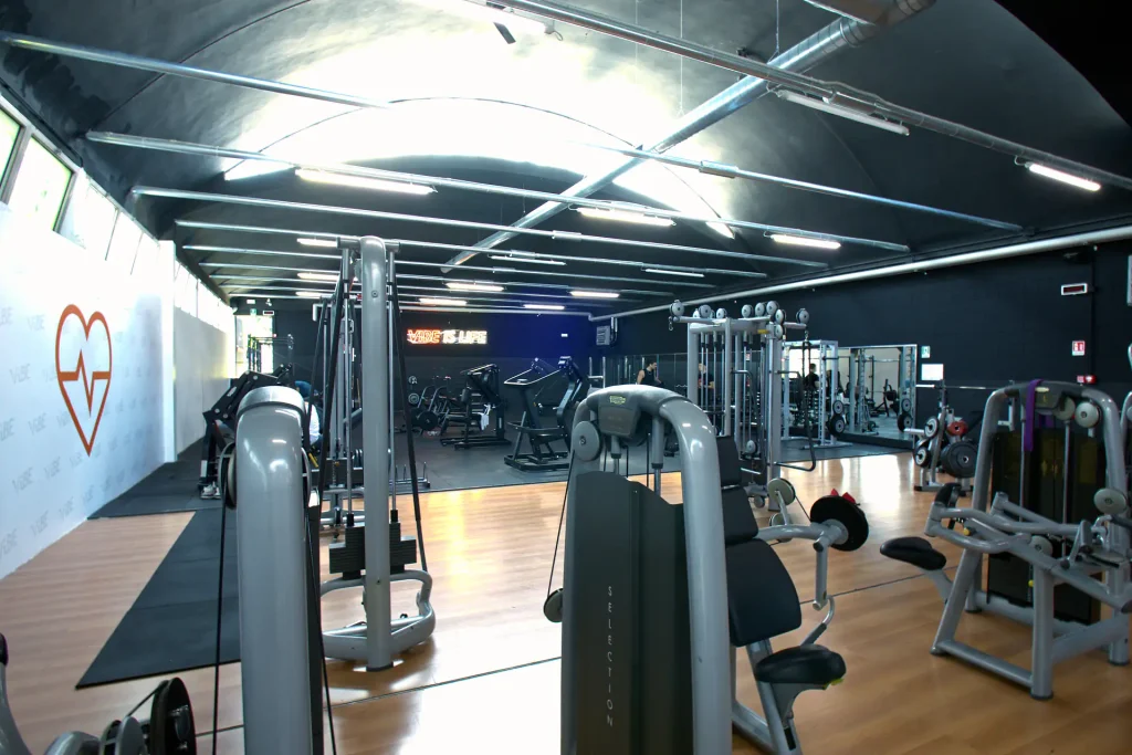 Vista laterale della sala attrezzi con macchinari Technogym e logo a forma di cuore con battito cardiaco sulla parete di ViBE Fitness.