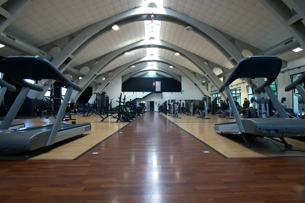 Corridoio centrale della sala attrezzi di ViBE Fitness Barlassina sotto la struttura ad archi in cemento, con tapis roulant Technogym Excite in primo piano.