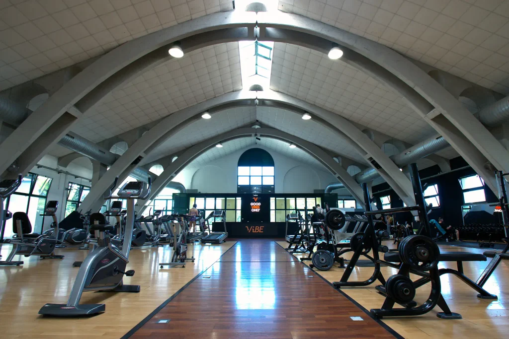 Vista frontale della sala pesi di ViBE Fitness Barlassina lungo il camminamento centrale in parquet scuro, con macchinari per l'allenamento ai lati.