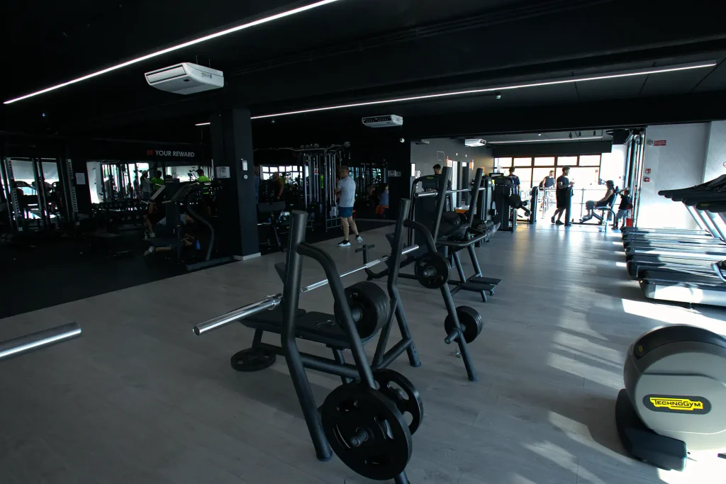 Prospettiva della sala attrezzi di ViBE Fitness Lentate con panche per bilancieri in primo piano e fila di tapis roulant Technogym sulla destra.