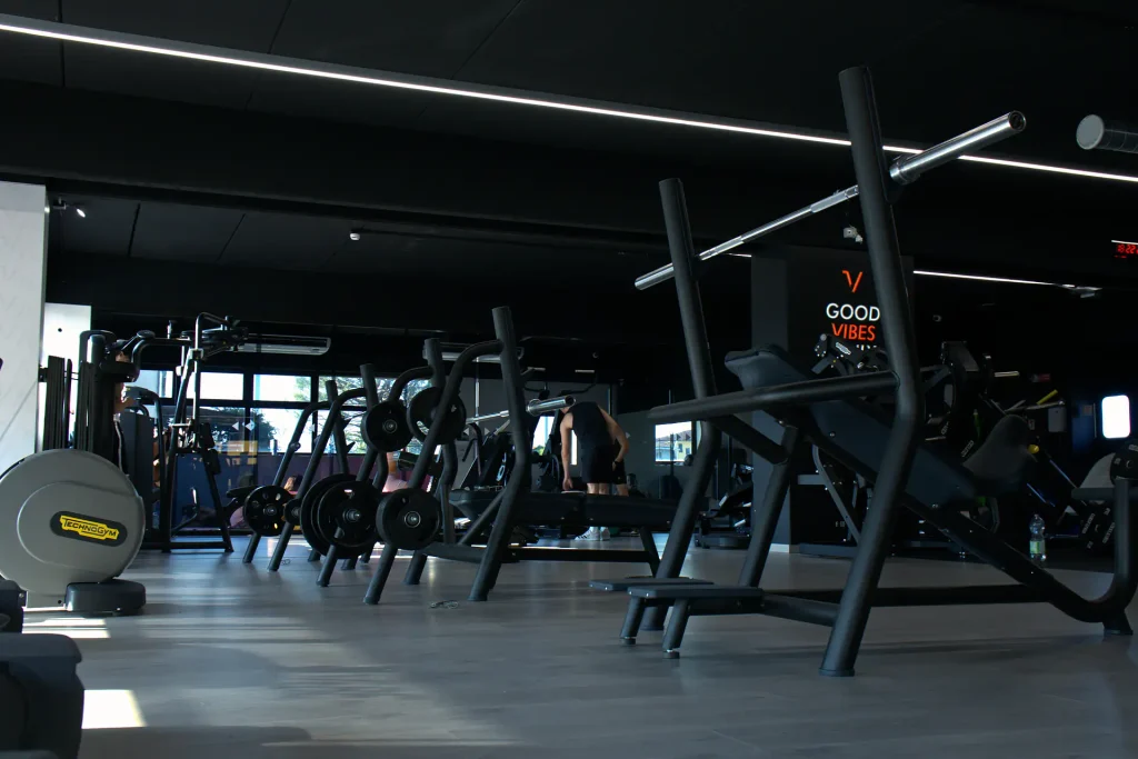 Inquadratura ad angolo della sala pesi di ViBE Fitness Lentate con postazioni per bilancieri, rack e grafiche motivazionali a parete.