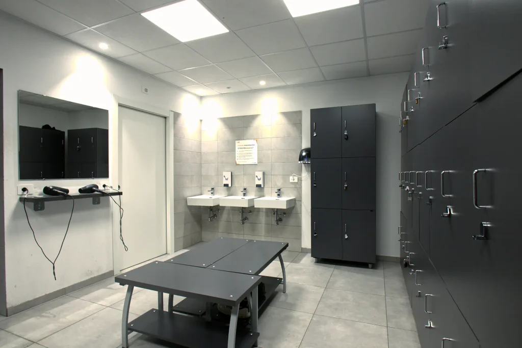 Interno dello spogliatoio di ViBE Fitness Lentate con armadietti grigio scuro, lavabi sospesi, specchio con phon e panca centrale.