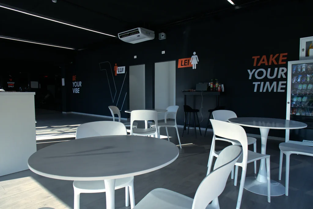 Zona lounge di ViBE Fitness Lentate con tavolini rotondi bianchi, sedie moderne, distributori automatici di bevande e scritte grafiche sulle pareti nere.