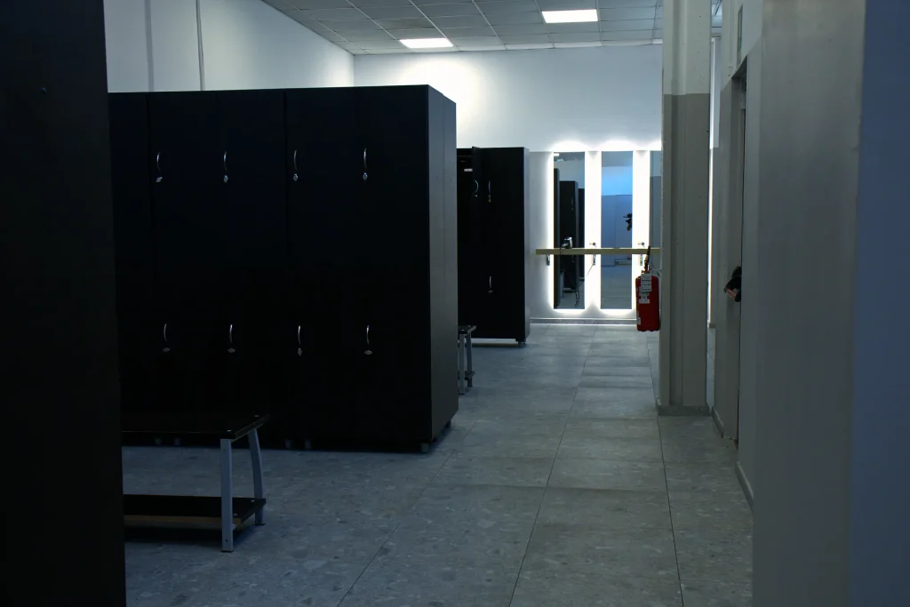 Una fila di armadietti neri a figura intera lungo un muro, con una panchina scura di fronte. In fondo, specchi con luci a LED e un estintore rosso sono visibili su un pavimento di piastrelle grigie.