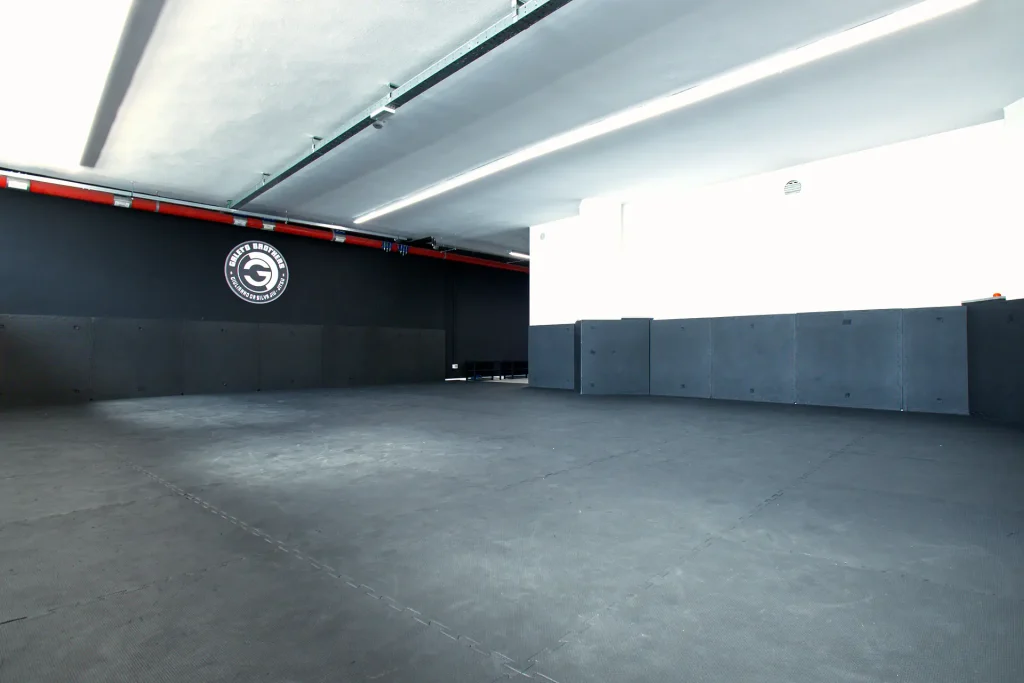 Una grande sala vuota dedicata alle arti marziali con pavimento e pareti rivestiti di tappetini neri imbottiti. Sulla parete di fondo nera è visibile il logo circolare 