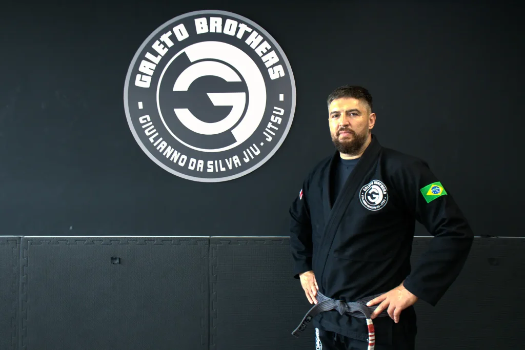 Un istruttore di Brazilian Jiu-Jitsu in piedi con un kimono nero e cintura nera davanti a un muro nero con il logo 