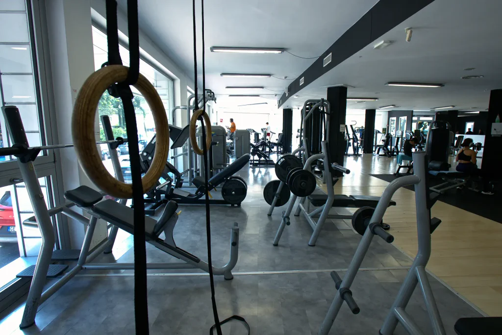 Una prospettiva di diverse macchine da palestra, tra cui una leg press in primo piano e altre attrezzature da cardio e pesi sullo sfondo, all'interno di una sala luminosa con grandi finestre.