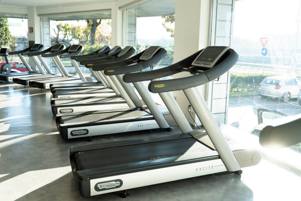 Una serie di tapis roulant Technogym moderni allineati lungo una parete con grandi finestre che mostrano l'esterno. La sala è spaziosa con pavimenti grigi.