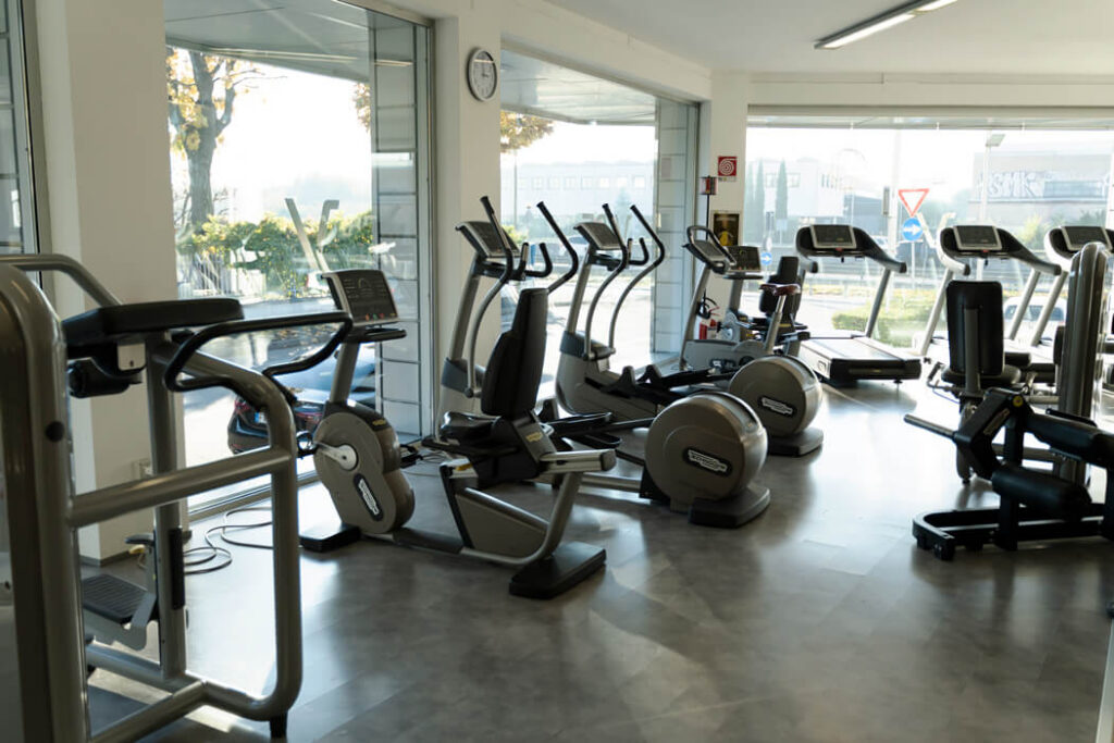 Zona cardio di Vibe Fitness Club Varedo illuminata da vetrate naturali, attrezzata con cyclette e macchine ellittiche Technogym.