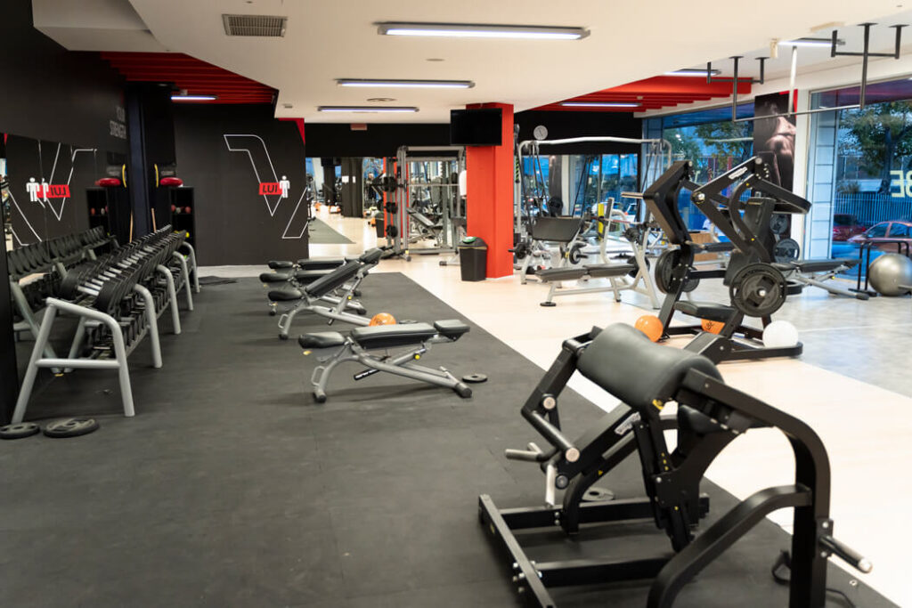 Area allenamento pesi liberi con pavimentazione gommata nera, rastrelliera completa di manubri, panchine regolabili e macchine pesi Technogym.